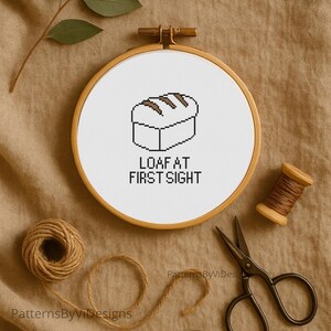 May include: A cross-stitch embroidery hoop with a bread loaf design and the text "LOAF AT FIRST SIGHT". The embroidery is set against a neutral background, with crafting supplies like scissors and thread spools.