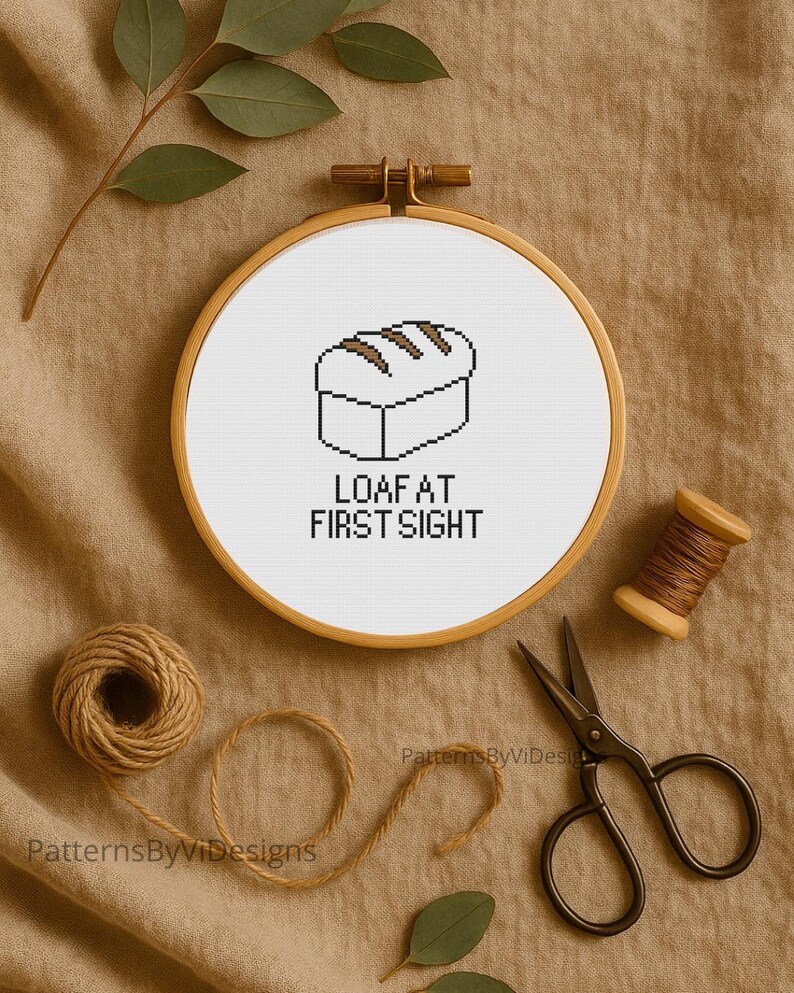 May include: A cross-stitch embroidery hoop with a bread loaf design and the text "LOAF AT FIRST SIGHT". The embroidery is set against a neutral background, with crafting supplies like scissors and thread spools.