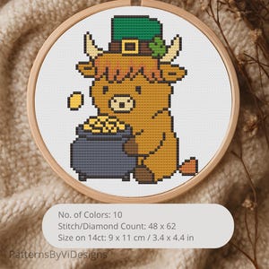 May include: A cross-stitch design of a Highland cow wearing a green leprechaun hat and holding a pot of gold. The design is in a wooden embroidery hoop. The image includes the text "No. of Colors: 10", "Stitch/Diamond Count: 48 x 62", and "Size on 14ct: 3.4 x 4.4 in".
