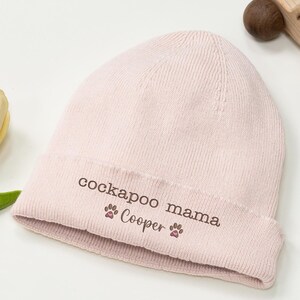 May include: A light pink beanie with the text "cockapoo mama" and "Cooper" printed on the folded brim. The beanie is next to a wooden toy car and a yellow and white flower.