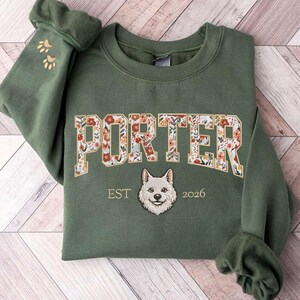 May include: Green sweatshirt with the name "PORTER" in floral letters. Below the name is a dog's face and the text "EST 2026". The left sleeve cuff has paw prints.