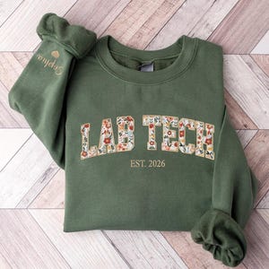 May include: An olive green sweatshirt with the words "LAB TECH" in a floral design across the chest. The text "EST. 2026" is below the main text. The sleeve cuff has the name "Sophia" embroidered with a small heart.