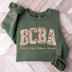 May include: An olive green sweatshirt with the letters "BCBA" in a floral design. Below the letters is the text "Board Certified Behavior Analyst". The sleeve has the name "Sophia" embroidered in gold.