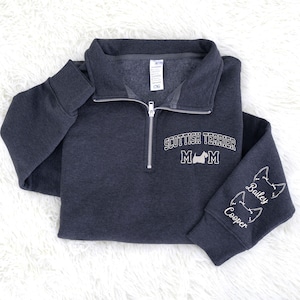 May include: Dark gray sweatshirt with a quarter-zip closure. The text "SCOTTISH TERRIER MOM" is printed on the chest, along with a dog silhouette. The sleeve features dog silhouettes and the names "Bailey" and "Cooper".
