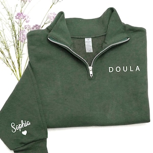 K&ouml;nnte beinhalten: Ein dunkelwaldgr&uuml;nes Sweatshirt mit Viertelrei&szlig;verschluss aus silberfarbenem Metall. Das Wort "DOULA" ist in Wei&szlig; auf der rechten Brust aufgedruckt, und der Name "Sophia" mit einem kleinen Herzsymbol befindet sich in Wei&szlig; am linken &Auml;rmelb&uuml;ndchen.