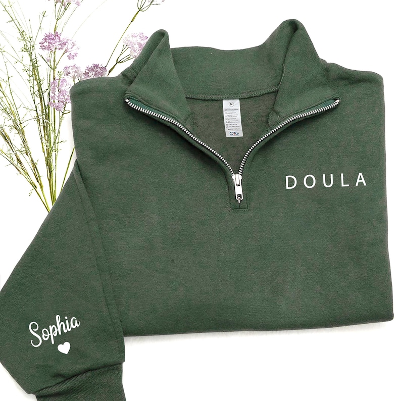 K&ouml;nnte beinhalten: Ein dunkelwaldgr&uuml;nes Sweatshirt mit Viertelrei&szlig;verschluss aus silberfarbenem Metall. Das Wort "DOULA" ist in Wei&szlig; auf der rechten Brust aufgedruckt, und der Name "Sophia" mit einem kleinen Herzsymbol befindet sich in Wei&szlig; am linken &Auml;rmelb&uuml;ndchen.