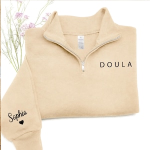 K&ouml;nnte beinhalten: Ein beigefarbenes Sweatshirt mit kurzem Rei&szlig;verschluss und dem Wort DOULA in schwarzem Druck auf der Brust. Auf dem &Auml;rmel steht der Name Sophia in schwarzer Schreibschrift &uuml;ber einem kleinen schwarzen Herzsymbol und einem silbernen Rei&szlig;verschluss.