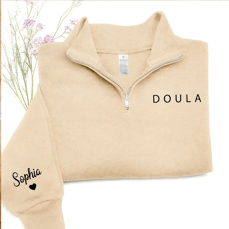 K&ouml;nnte beinhalten: Ein beigefarbenes Sweatshirt mit kurzem Rei&szlig;verschluss und dem Wort DOULA in schwarzem Druck auf der Brust. Auf dem &Auml;rmel steht der Name Sophia in schwarzer Schreibschrift &uuml;ber einem kleinen schwarzen Herzsymbol und einem silbernen Rei&szlig;verschluss.