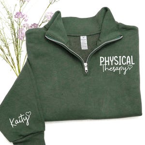 May include: A dark green quarter-zip sweatshirt with the words "Physical Therapy" in white script on the front and "Kaity" on the left sleeve. The sweatshirt has a silver zipper and a small white tag.