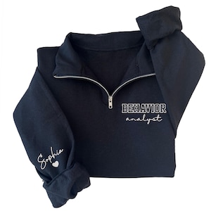 May include: Black quarter-zip sweatshirt with the text "BEHAVIOR analyst" in white. The left sleeve has the name "Sophia" and a heart symbol in white. The sweatshirt has a silver zipper.
