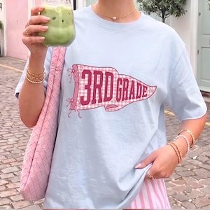 May include: A light blue short-sleeved t-shirt featuring a pink and white gingham pennant graphic with the text 3RD GRADE in bold dark pink letters. The design includes small star accents and decorative pink ribbons tied at the pennant's edge.