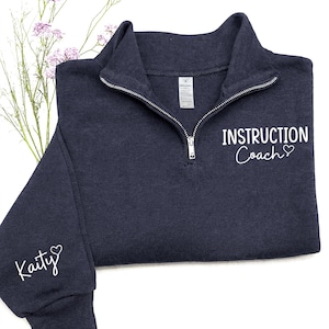 May include: A navy blue quarter-zip sweatshirt with the words "Instruction Coach" and a heart symbol printed in white. The sleeve features the name "Kaity" with a heart. The sweatshirt has a silver zipper and a tag.