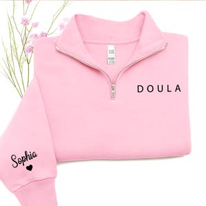 K&ouml;nnte beinhalten: Ein hellrosa Sweatshirt mit kurzem Rei&szlig;verschluss und dem Wort DOULA in schwarzem Druck auf der Brust. Auf dem linken &Auml;rmel steht der Name Sophia in schwarzer Schreibschrift, gefolgt von einem kleinen schwarzen Herzsymbol. Silberner Rei&szlig;verschluss.