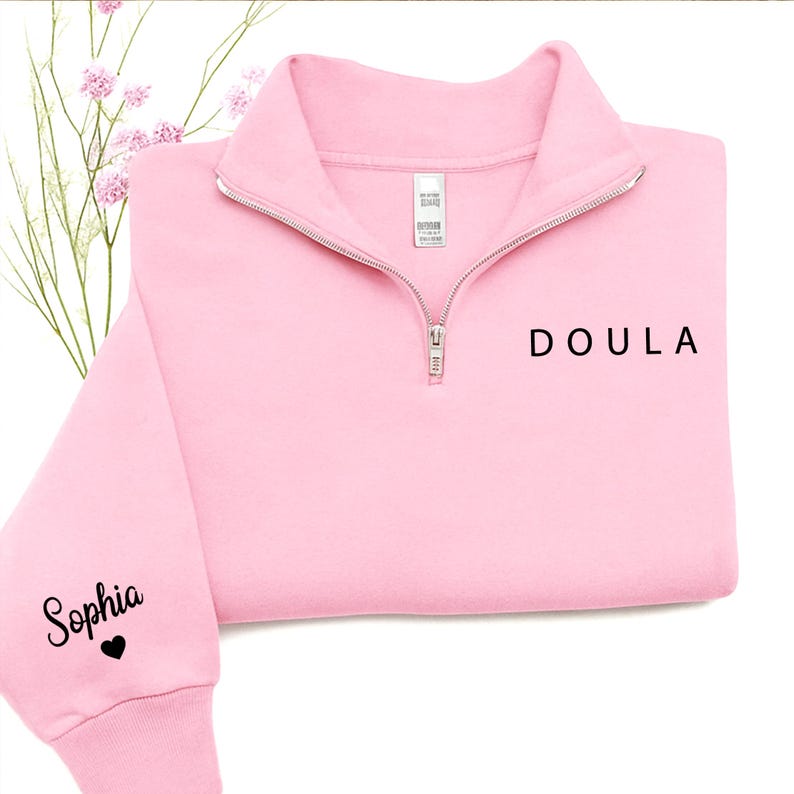 K&ouml;nnte beinhalten: Ein hellrosa Sweatshirt mit kurzem Rei&szlig;verschluss und dem Wort DOULA in schwarzem Druck auf der Brust. Auf dem linken &Auml;rmel steht der Name Sophia in schwarzer Schreibschrift, gefolgt von einem kleinen schwarzen Herzsymbol. Silberner Rei&szlig;verschluss.