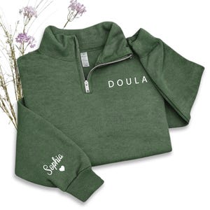 K&ouml;nnte beinhalten: Ein dunkelolivgr&uuml;nes Sweatshirt mit kurzem Rei&szlig;verschluss in Silberoptik. Auf der linken Brust ist das Wort DOULA in Wei&szlig; aufgedruckt. Der Name Sophia ist in wei&szlig;er Schreibschrift auf dem &Auml;rmel zu sehen, erg&auml;nzt durch ein kleines Herz.