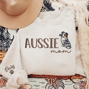 May include: Cream-colored sweatshirt with the embroidered text "AUSSIE mom" and a detailed illustration of an Australian Shepherd dog holding a flower. The left sleeve has the name "Daisy" and a paw print. A tan hat is visible in the upper left corner.