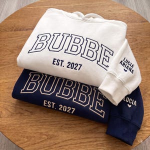 May include: A white hoodie with dark blue text reading BUBBE EST. 2027 and LUCIA ARIANA with a heart on the sleeve, stacked on a dark blue hoodie with light text reading BUBBE EST. 2027 and LUCIA with a heart on the sleeve, both featuring ribbed cuffs.
