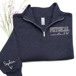 May include: A navy blue quarter-zip sweatshirt with the words "PHYSICAL assistant" embroidered in white. The name "Sophia" is embroidered on the sleeve. The sweatshirt has a silver zipper and a small white tag.