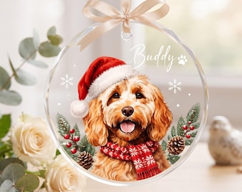 May include: A clear circular acrylic ornament featuring a golden-brown dog wearing a red Santa hat and a red patterned scarf. The name Buddy is printed in white script above the dog, accompanied by a paw print, snowflakes, pine, and berries.