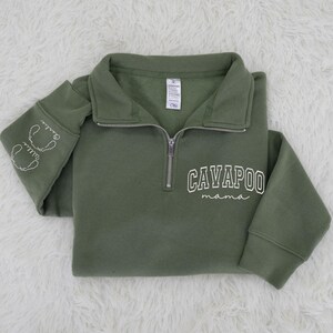 May include: Olive green Cavapoo Mama sweatshirt with a quarter-zip collar. The sweatshirt features the words "CAVAPOO mama" in white script on the front and embroidered dog faces with names on the left sleeve. The sweatshirt is folded on a white, fluffy surface.