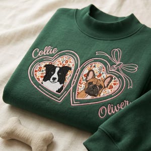 May include: A dark green mock neck sweatshirt featuring an embroidered design of two heart-shaped lockets. The left locket contains a black and white Border Collie, and the right contains a fawn French Bulldog, both with floral backgrounds.