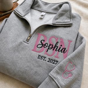 May include: A heather gray quarter-zip sweatshirt featuring pink embroidered lettering that reads BSN with the name Sophia in black script overlaid. Below the name is the text EST. 2027 in black, and a pink stethoscope icon on the sleeve cuff.