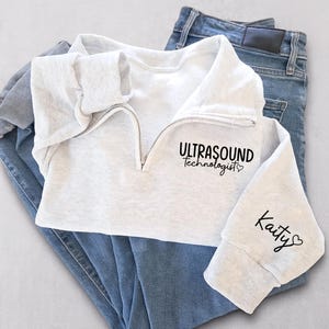 May include: A light gray sweatshirt with a quarter-zip neckline, the words "ULTRASOUND Technologist" printed on the front, and the name "Kaity" on the sleeve. The sweatshirt is paired with blue jeans.