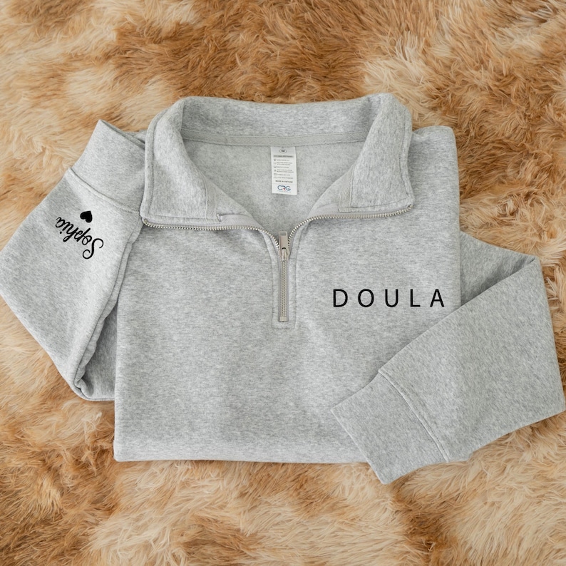 K&ouml;nnte beinhalten: Ein hellgraues Sweatshirt mit kurzem Rei&szlig;verschluss und dem Wort DOULA in schwarzem Druck auf der linken Brust. Auf dem linken &Auml;rmel steht der Name Sophia in schwarzer Schreibschrift, erg&auml;nzt durch ein kleines schwarzes Herzsymbol.