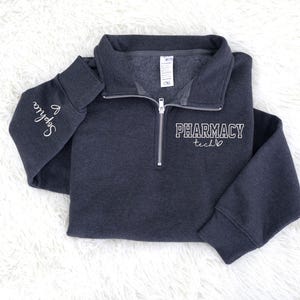 May include: A dark gray quarter-zip sweatshirt with the words "PHARMACY tech" in white script on the front. The left sleeve has the name "Sophia" embroidered in white. The sweatshirt is lying on a white, fluffy surface.