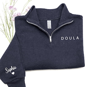 K&ouml;nnte beinhalten: Ein dunkelblaues Sweatshirt mit kurzem Rei&szlig;verschluss und dem Wort DOULA in wei&szlig;en Gro&szlig;buchstaben auf der Brust. Auf dem linken &Auml;rmel steht der Name Sophia in wei&szlig;er Schreibschrift &uuml;ber einem kleinen wei&szlig;en Herzsymbol.