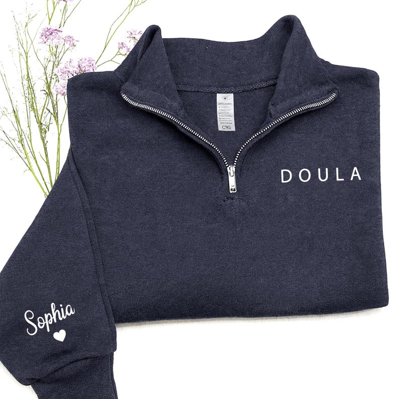 K&ouml;nnte beinhalten: Ein dunkelblaues Sweatshirt mit kurzem Rei&szlig;verschluss und dem Wort DOULA in wei&szlig;en Gro&szlig;buchstaben auf der Brust. Auf dem linken &Auml;rmel steht der Name Sophia in wei&szlig;er Schreibschrift &uuml;ber einem kleinen wei&szlig;en Herzsymbol.
