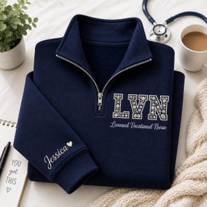 May include: A navy blue quarter-zip sweatshirt featuring the embroidered letters LVN filled with a white daisy pattern. Below the letters, the text reads Licensed Vocational Nurse. The sleeve is embroidered with the name Jessica and a small heart.
