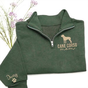 May include: A dark green quarter-zip sweatshirt with a silver zip. The sweatshirt has the words "CANE CORSO mom" and a dog silhouette embroidered in light brown. The name "Daisy" is embroidered on the sleeve.