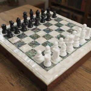 May include: A chess set with black and white pieces on a checkered board. The board has a dark wood frame and a decorative mother-of-pearl inlay. The chess pieces are arranged in their starting positions. The board measures approximately 30 cm x 30 cm.
