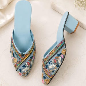 May include: A pair of light blue embroidered mules. The open-toe shoes feature a block heel and intricate floral embroidery in shades of pink, yellow, and green. The heels are approximately 7.6 cm high.