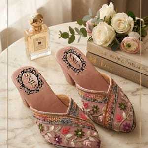 May include: A pair of pink embroidered mules with a block heel. The shoes feature floral embroidery in shades of green, red, and purple. The insole has the text "SNJLA" in a decorative oval. A bottle of perfume and a bouquet of flowers are in the background.