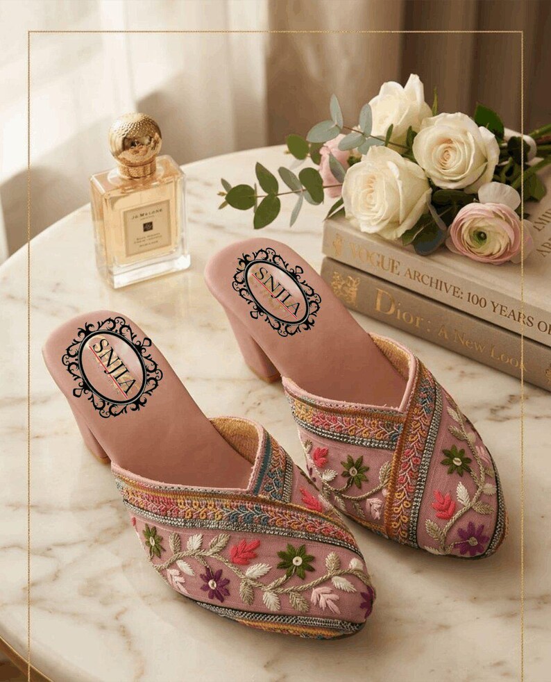 May include: A pair of pink embroidered mules with a block heel. The shoes feature floral embroidery in shades of green, red, and purple. The insole has the text "SNJLA" in a decorative oval. A bottle of perfume and a bouquet of flowers are in the background.