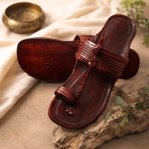 Op de afbeelding: Een paar donkerbruine leren sandalen met een teenlusontwerp, een gevlochten band die de teenlus verbindt met de brede wreefband, en ingewikkelde in reliëf gemaakte lineaire patronen langs de randen en banden van het schoeisel.