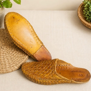 May include: A pair of tan leather slip-on shoes featuring a woven upper design. One shoe is positioned to show the smooth, solid leather sole, while the other displays the intricate braided texture of the top and the interior footbed.