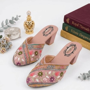 May include: A pair of pink embroidered mules with a block heel. The mules feature floral embroidery in various colors. The footbed has the text "SNJILA" in a decorative oval. The shoes are displayed with books and decorative items.