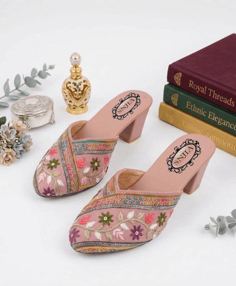 May include: A pair of pink embroidered mules with a block heel. The mules feature floral embroidery in various colors. The footbed has the text "SNJILA" in a decorative oval. The shoes are displayed with books and decorative items.