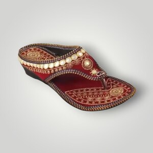 Może przedstawiać: Bordowa sandała z misternym złotym haftem i małymi lustrzanymi akcentami. Sandał ma klinowy obcas i pasek w stylu string&oacute;w. Projekt obejmuje szczeg&oacute;łowe wzory i zdobienia, prezentujące mieszankę element&oacute;w tradycyjnych i dekoracyjnych.