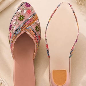 May include: A pair of pink embroidered slip-on shoes. The uppers are adorned with colorful floral embroidery. The soles are a light pink with a small, tan heel. The shoes are designed for comfort and style.