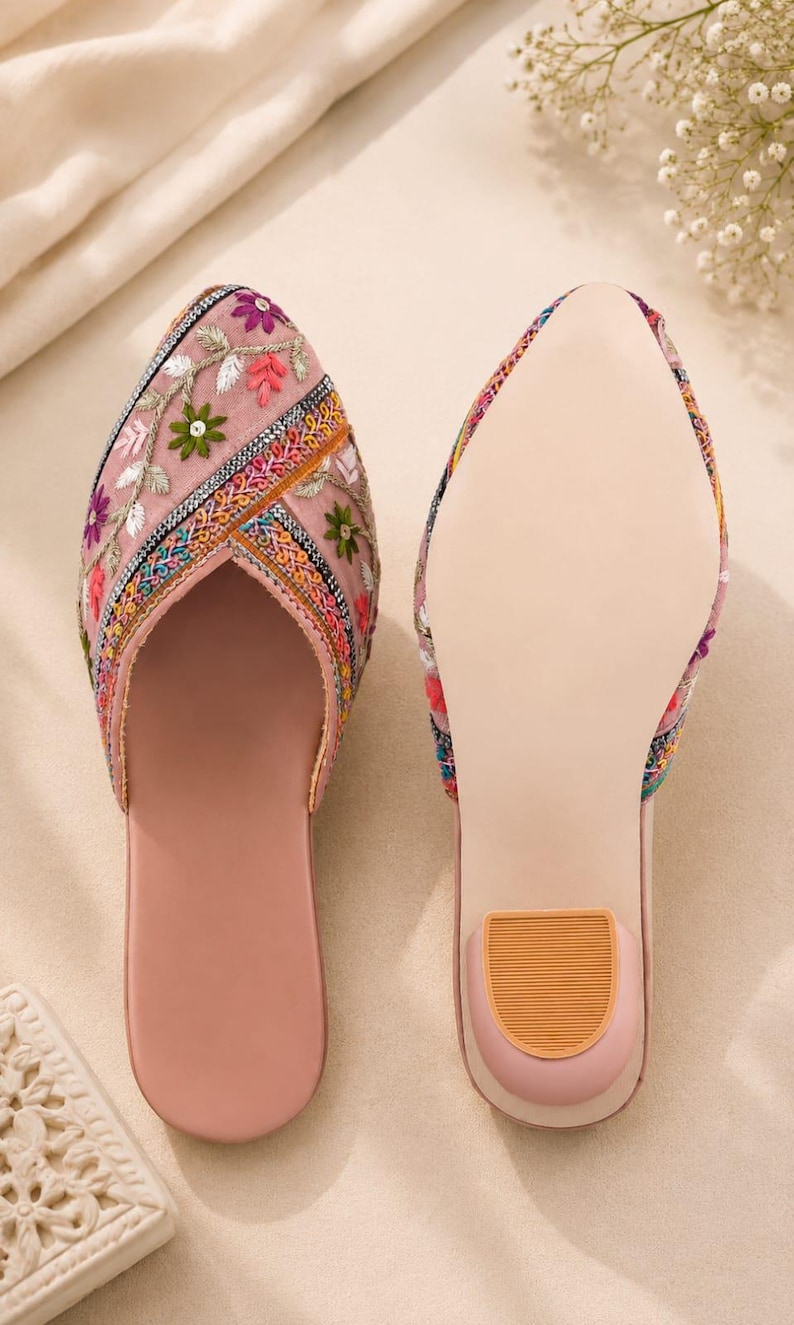 May include: A pair of pink embroidered slip-on shoes. The uppers are adorned with colorful floral embroidery. The soles are a light pink with a small, tan heel. The shoes are designed for comfort and style.