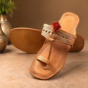 May include: A pair of tan leather sandals featuring a toe post, a wide strap with intricate embossed patterns, metallic thread detailing, and a small red tassel. The footbed is stamped with the text IND-KOLHAPURI LEATHER S.G. and the number 9.