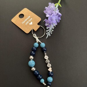 Può includere: Un portachiavi fatto a mano con perline blu e argento che compongono la parola "BEN" e un ciondolo a forma di cuore. Il portachiavi è attaccato a un'etichetta marrone con la scritta "Handmade with love". Un rametto di fiori viola è sullo sfondo.