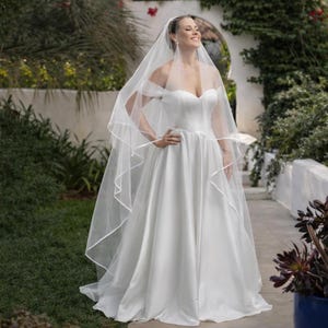 May include: A bride in a white satin wedding dress with a sweetheart neckline and a flowing skirt. She is wearing a sheer veil with ribbon trim. The photo is taken outdoors with greenery in the background.