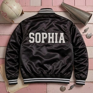 May include: A black satin varsity jacket featuring the name SOPHIA in large white block letters across the back. The jacket has a ribbed collar, cuffs, and hem with white stripes, long sleeves, and a button-up front closure design.