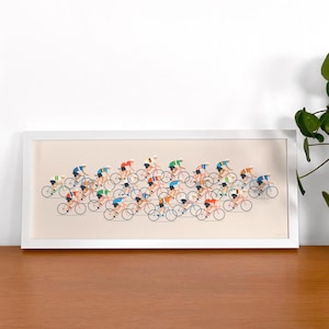 Op de afbeelding: Een ingelijste horizontale kunstdruk met een gestileerde illustratie van een grote groep wielrenners tijdens een race. De wielrenners dragen kleurrijke truien en witte helmen, afgebeeld in een minimalistische grafische stijl op een beige achtergrond.