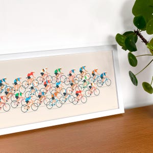 Op de afbeelding: Een ingelijste horizontale print met een gestileerde illustratie van een grote groep wielrenners in kleurrijke truien op fietsen. Het kunstwerk is geplaatst tegen een lichtbeige achtergrond en toont een peloton renners in diverse posities.
